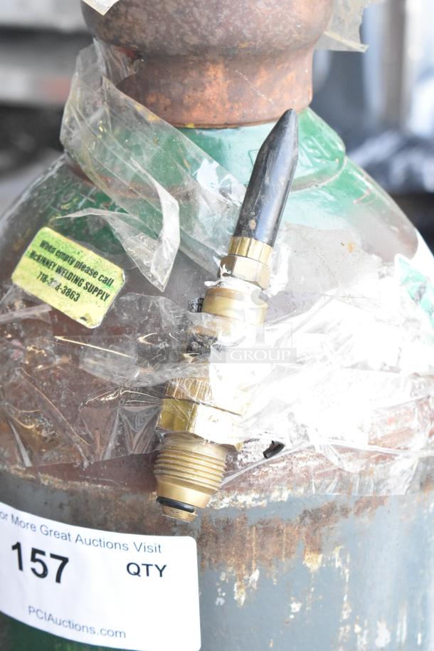 Rusty metal tank with fittings, partially wrapped in plastic. Label suggests McKinney Welding Supply contact when empty. Manual pickup required.