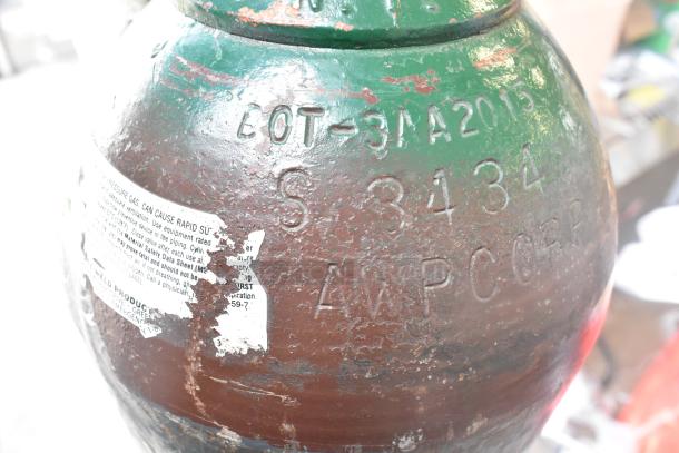 Large green and brown metal tank with engravings "BOT-31A2015 S 3434 AWP CORP." Peeling label warns of gas pressure.