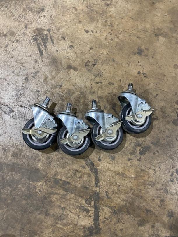 Four metal casters with rubber wheels on a wooden surface, featuring locking brakes and swivel mounts.