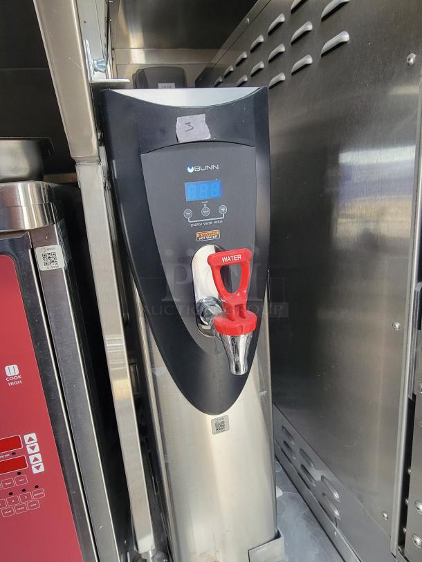 Bunn commercial hot water dispenser with red tap in a stainless steel mobile kitchen. Model details and settings shown.