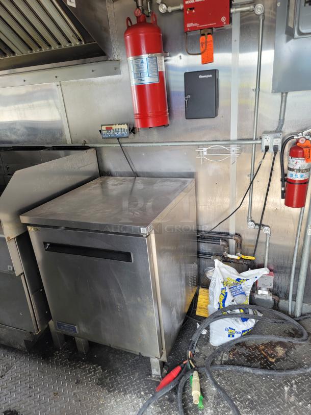 Interior of a Diamond Cargo mobile kitchen featuring stainless steel surfaces, commercial refrigerator, griddle, and fire safety equipment.