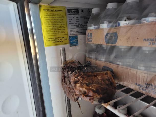 Interior of Arctic Air refrigeration unit with warning label about condenser coil inspection. Includes bottled water and decayed item.
