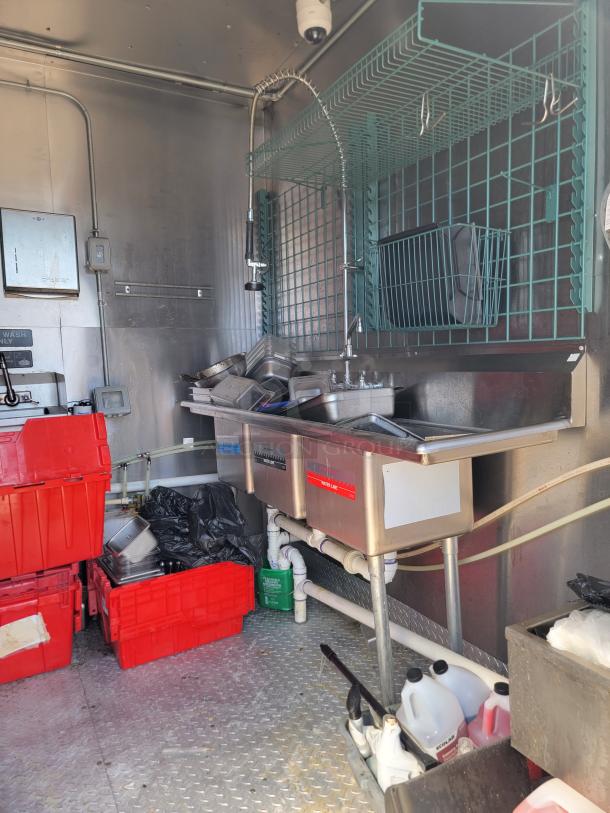 Interior of a 2021 Diamond Cargo mobile kitchen; features a stainless steel sink, industrial shelving, and red storage bins.
