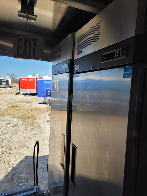 Interior view of a 2021 Rock Solid Cargo 28’ mobile kitchen showing a stainless steel commercial fridge, exit sign, and natural light.