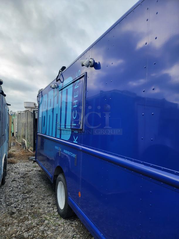 Blue food truck with "BRAND KITCHENS" label, side serving window, white wheels, and exterior lights. Weathered condition.