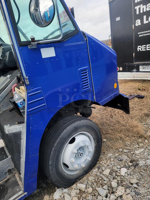 Blue delivery truck with visible front tire and open driver's side door. The truck is on a rocky surface. The condition is used.