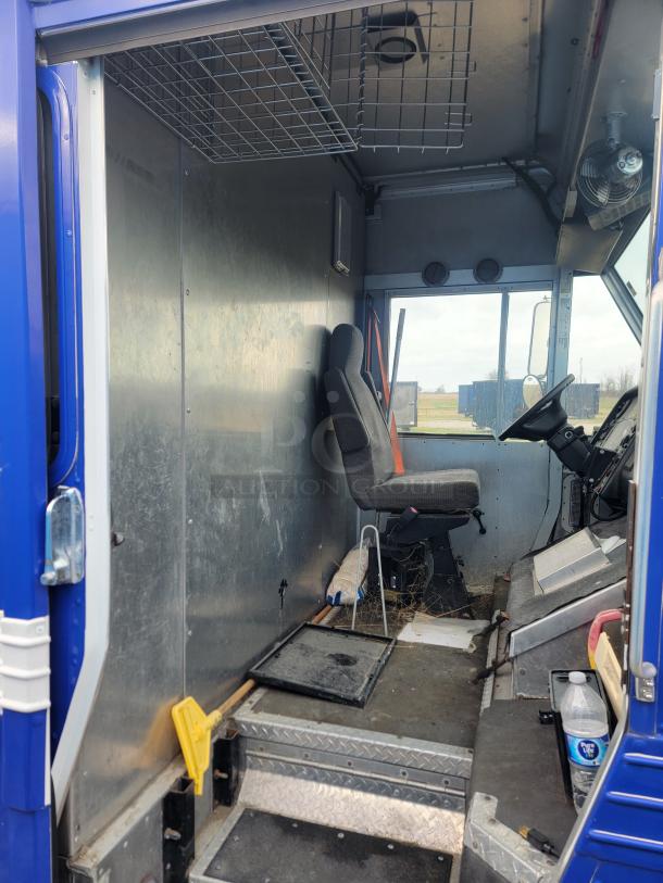 Interior of a blue delivery truck cabin showing a driver's seat, metal interior panels, wire storage shelf, and functional wear.