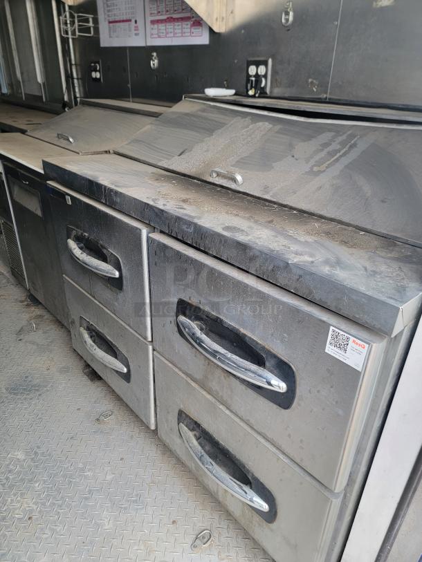 Stainless steel commercial kitchen prep station with multiple drawers, showing signs of use and dust accumulation. QR code label visible.