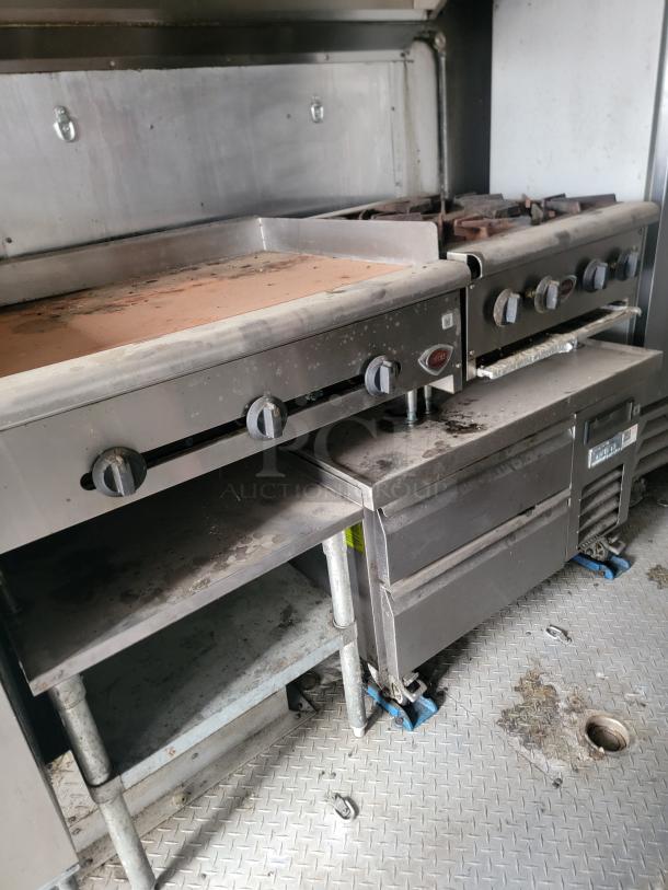 Commercial kitchen stove with griddle and multiple burners. Stainless steel, shows signs of use. Features gas knobs and utility space below.
