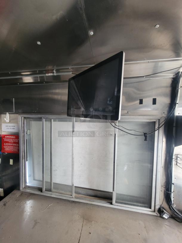 Wall-mounted flat-screen monitor in stainless steel setting, sliding glass doors below, power cables visible. Warnings posted on left.