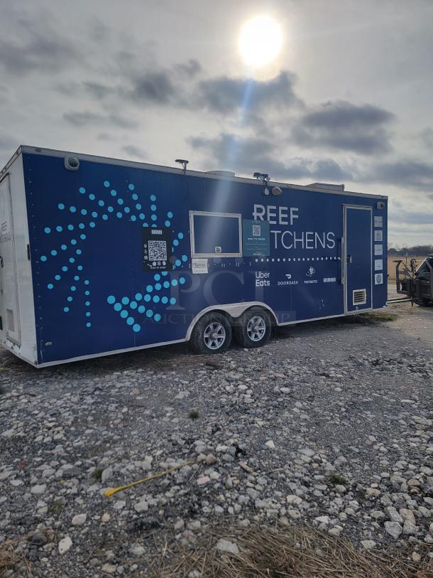 Food trailer with "REEF KITCHENS" branding, blue exterior, QR codes, delivery service logos, on gravel lot.