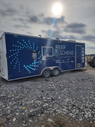 Food trailer with "REEF KITCHENS" branding, blue exterior, QR codes, delivery service logos, on gravel lot.