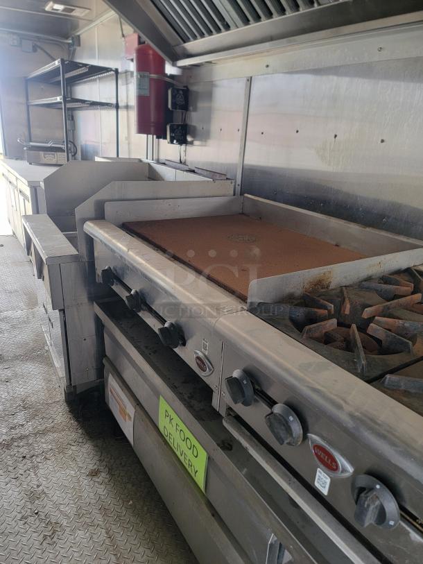 Commercial kitchen equipment with stainless steel range and grill. Visible brand: Wells. Includes shelving, exhaust hood, and fire extinguisher.