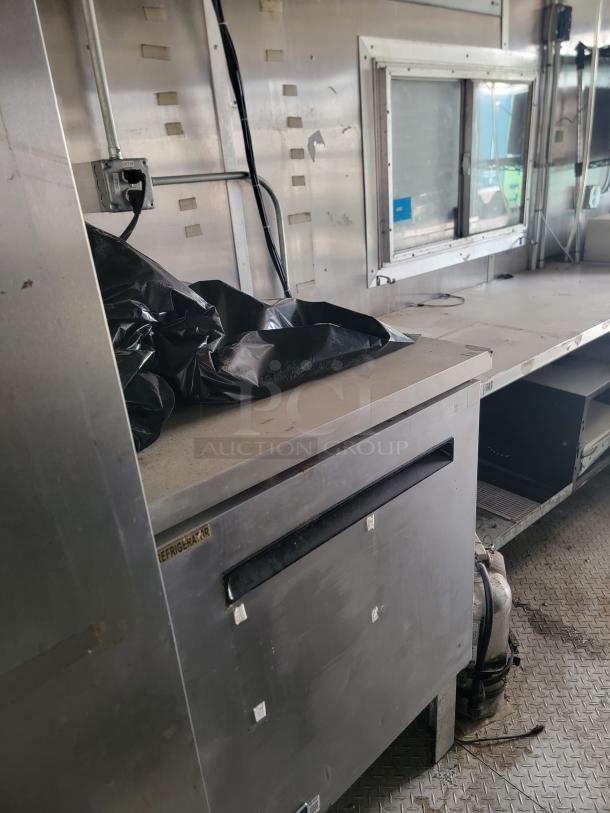 Industrial stainless steel refrigerator in a mobile unit, shows slight wear. Visible "Refrigerator" label. No other markings.