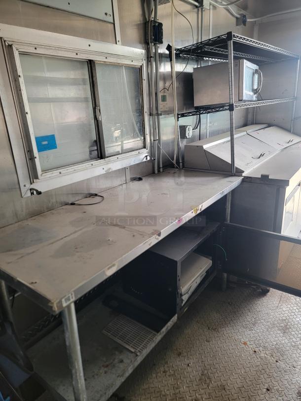 Commercial kitchen workspace with stainless steel shelves, microwave, and service window. Signs of wear visible.