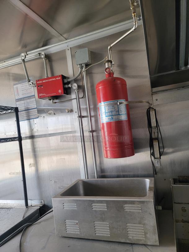 Commercial kitchen fire suppression system with red control box and extinguisher. Metal sink below, stainless steel walls.