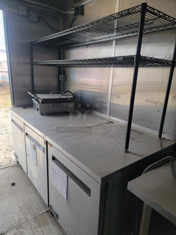 Commercial kitchen setup with Uline metal shelves and a countertop grill. Features under-counter storage units with visible wear.