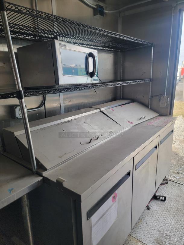 Commercial stainless steel prep table with compartments, upper shelving, and microwave. Good condition, ideal for food service.
