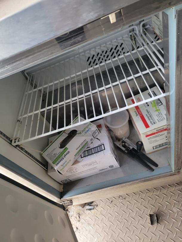 Commercial refrigerator interior showing wire shelves, vinyl gloves box, a plastic container, and utensils. Used condition.