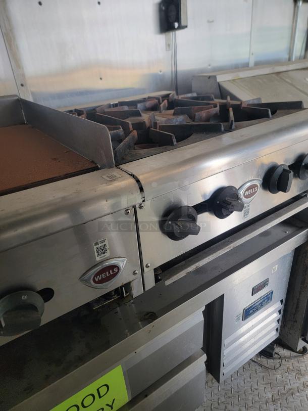 Commercial stove with Wells brand logo, four gas burners, stainless steel finish, visible control knobs, used condition.