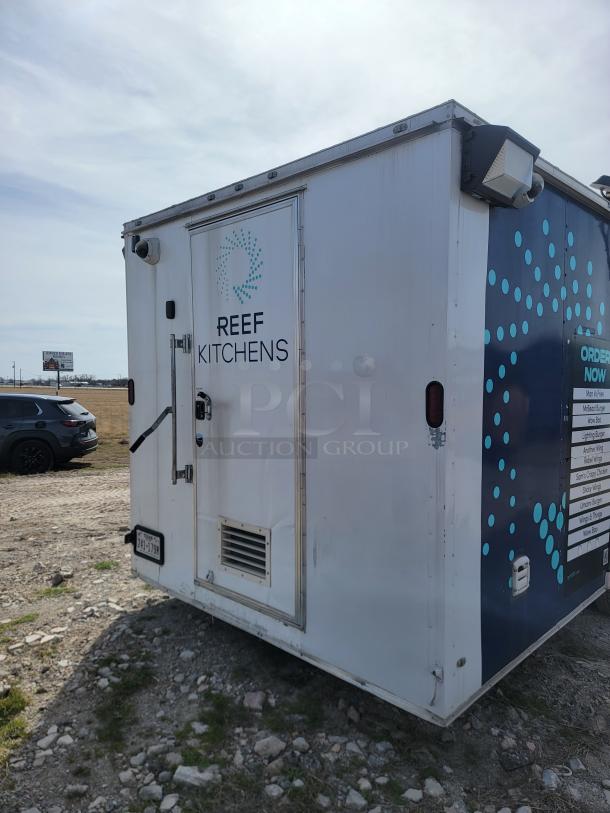 Mobile kitchen unit with "REEF KITCHENS" logo, white panel with ventilation hatch, and external lighting fixtures.