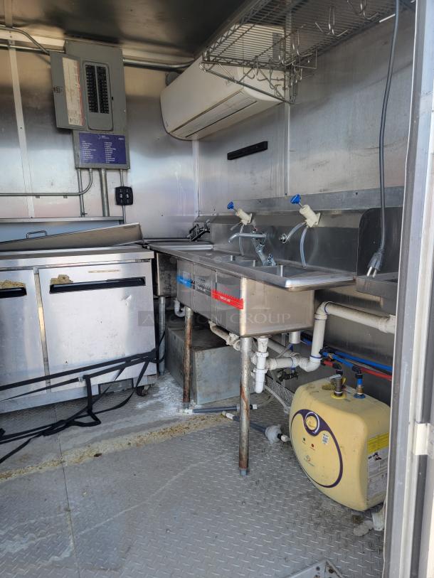 Interior of a mobile kitchen unit with stainless steel countertop, two sinks, plumbing, and an air conditioning unit.