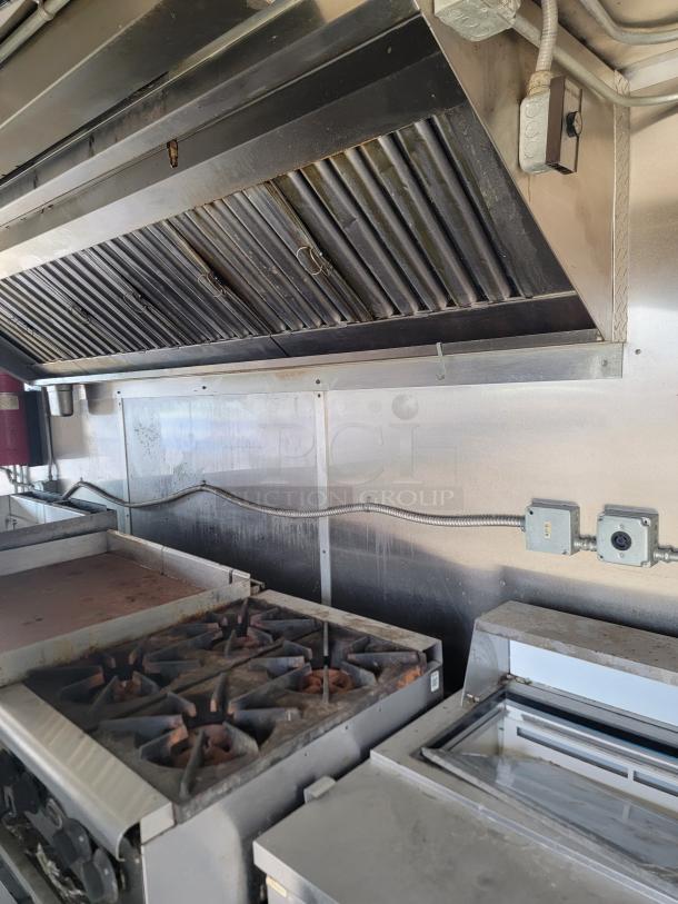 Commercial kitchen setup with six-burner gas stove, griddle, and overhead exhaust hood. Slight wear visible.