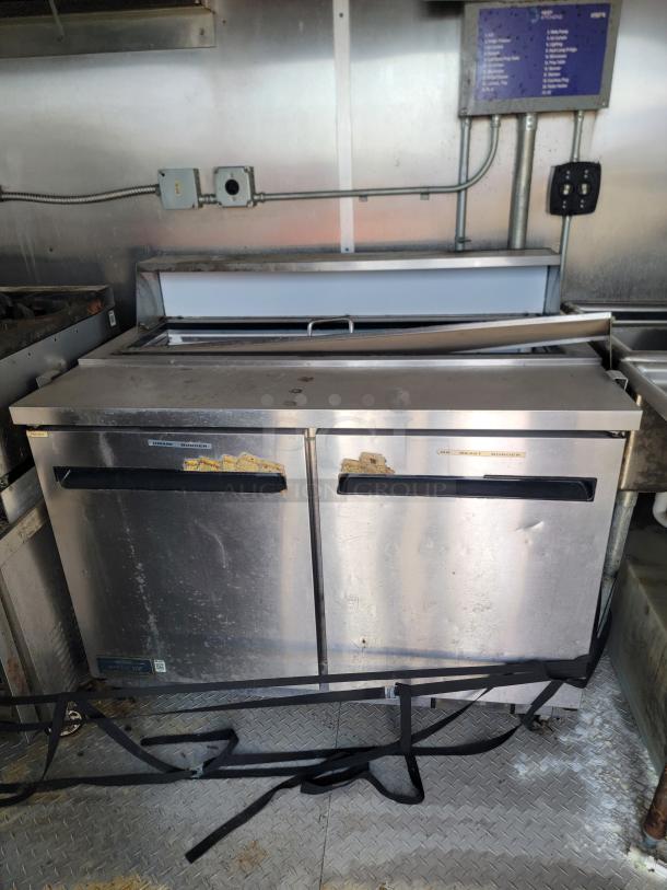 Commercial stainless steel under-counter refrigerator with visible wear and rust spots. Dual doors, strap at the base, located in a metal-walled kitchen.