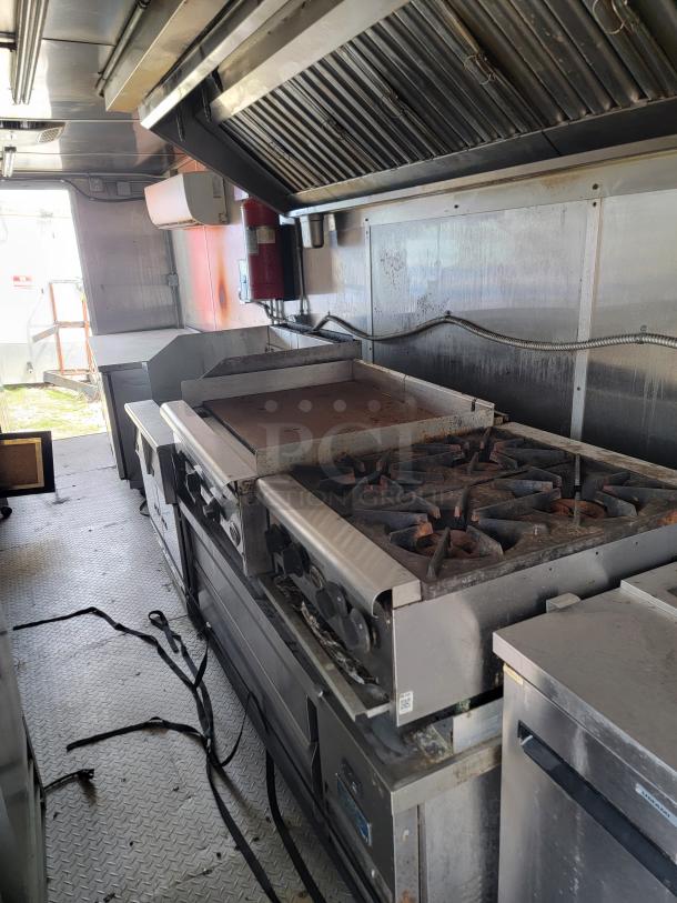 Commercial kitchen equipment in a mobile food truck, featuring a gas stove with multiple burners and a flat-top griddle. Condition shows visible wear and use.