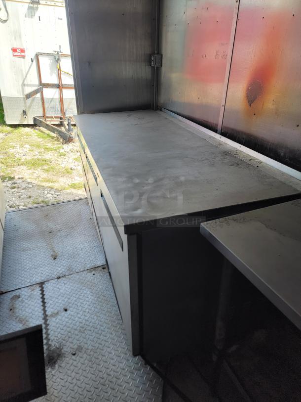 Stainless steel commercial workbench in used condition with visible wear, set on a diamond plate floor in an industrial setting.