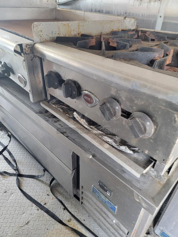Wells commercial gas range with multiple burners, visible wear and rust, includes control knobs and manufacturer's logo.