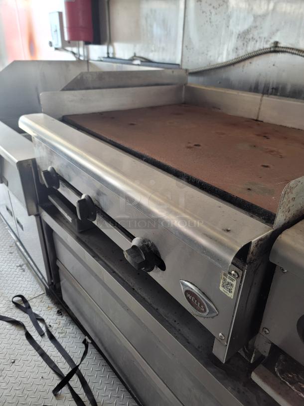 Wells commercial griddle, stainless steel, shows wear, two control knobs, branded label visible, kitchen setting.