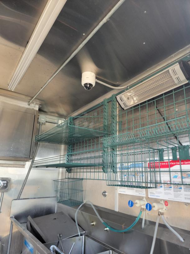 Interior of a food truck with metal shelves and baskets, a ceiling security camera, fluorescent lighting, and a stainless steel sink with spray hose.
