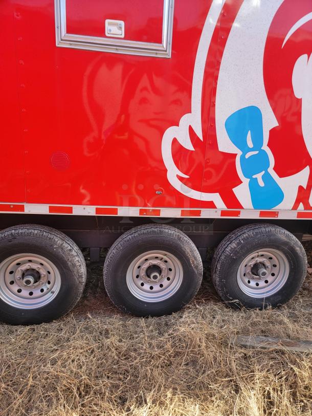 Red trailer with iconic fast-food graphic, triple axel polished wheels, and a small service door, on dry grass.