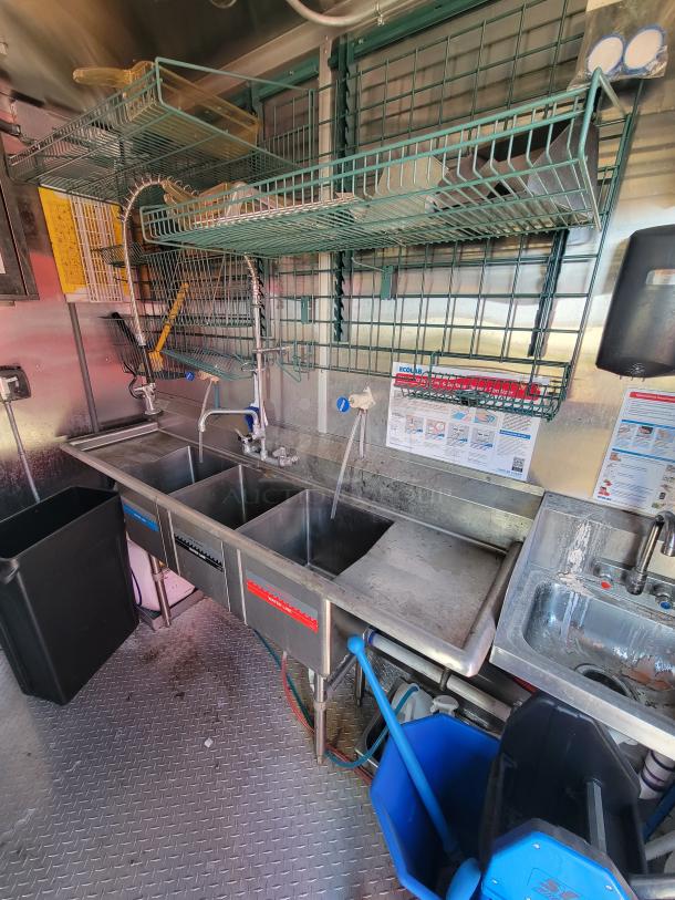 Stainless steel triple sink setup in commercial kitchen, includes two faucets, drying rack above. Clean condition.