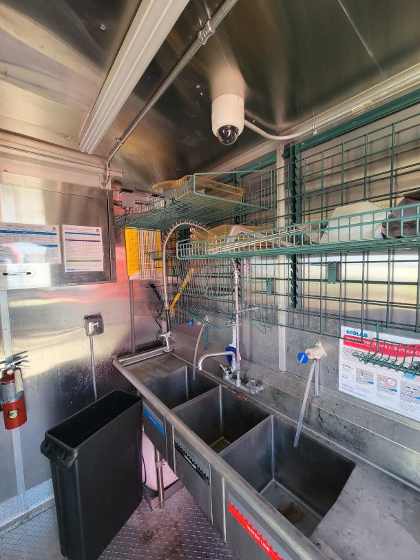 Commercial stainless steel three-compartment sink with attached faucet and overhead wire shelves. Features safety signs and fire extinguisher.