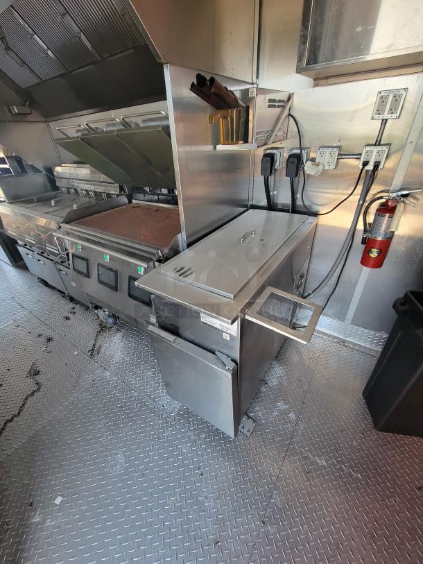Commercial kitchen equipment on diamond plate floor, featuring a flat-top grill, stainless steel surfaces, and side storage drawer.