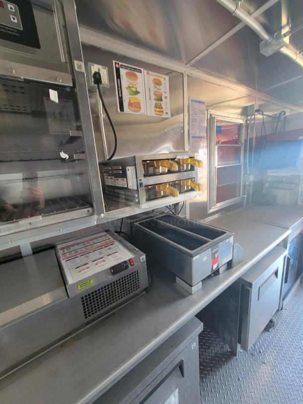 Commercial kitchen equipment inside a food truck, featuring stainless steel counters, storage units, and food preparation appliances.
