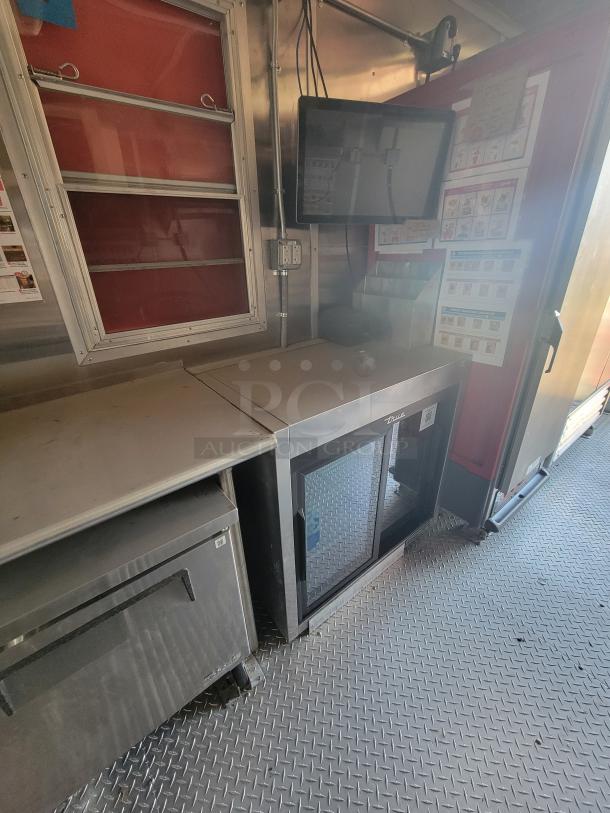 Commercial kitchen equipment in a metal food truck interior, includes a stainless steel counter, under-counter refrigerator, and mounted display. Labels indicate menu items. Clean and well-maintained.