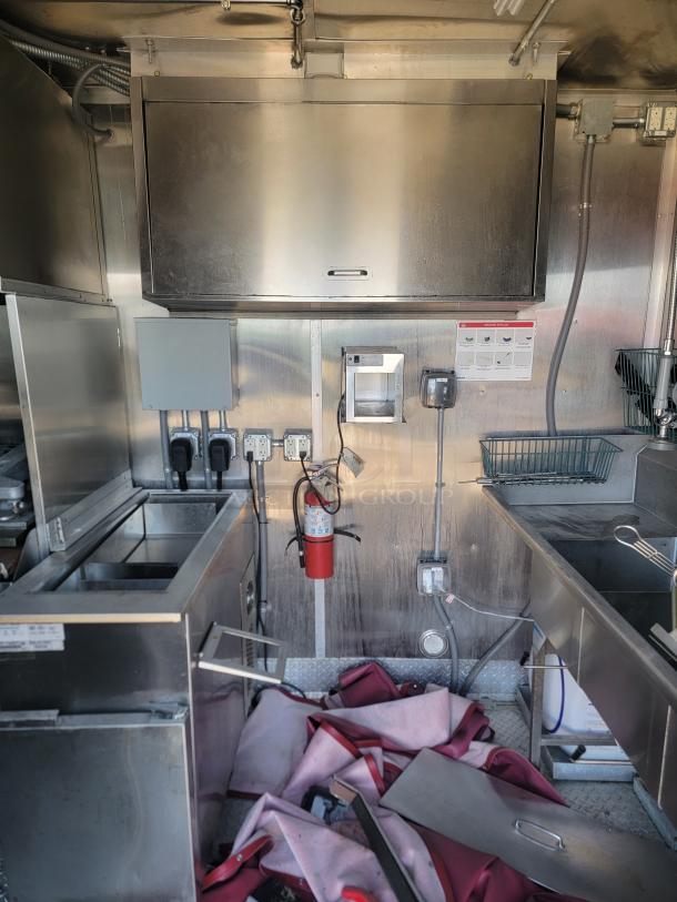 Commercial kitchen setup with stainless steel appliances, including a large vent hood and fryer. Fire extinguisher and electrical panel visible.