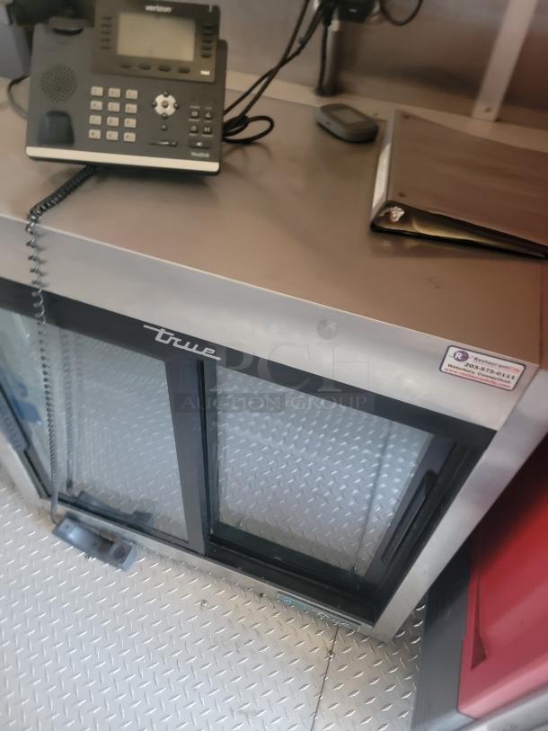 True commercial refrigerator with glass double doors, black frame, and silver top. Located on a diamond plate floor. Good condition.