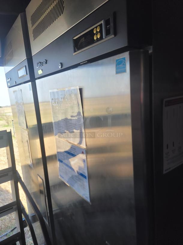 Commercial stainless steel refrigerator with digital controls and vents. Visible stickers and paper on the front. Good condition.
