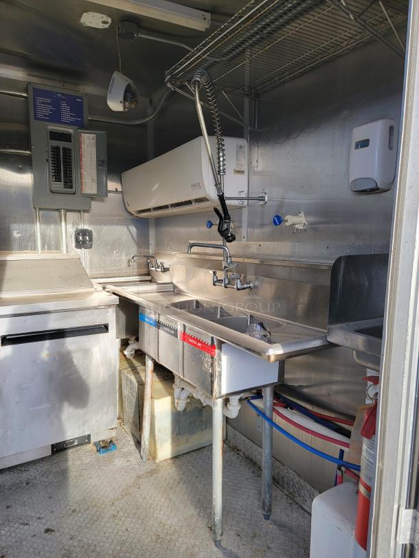 Commercial kitchen sink setup with two basins, stainless steel, mounted hoses. Contains a fire extinguisher and electrical panel.