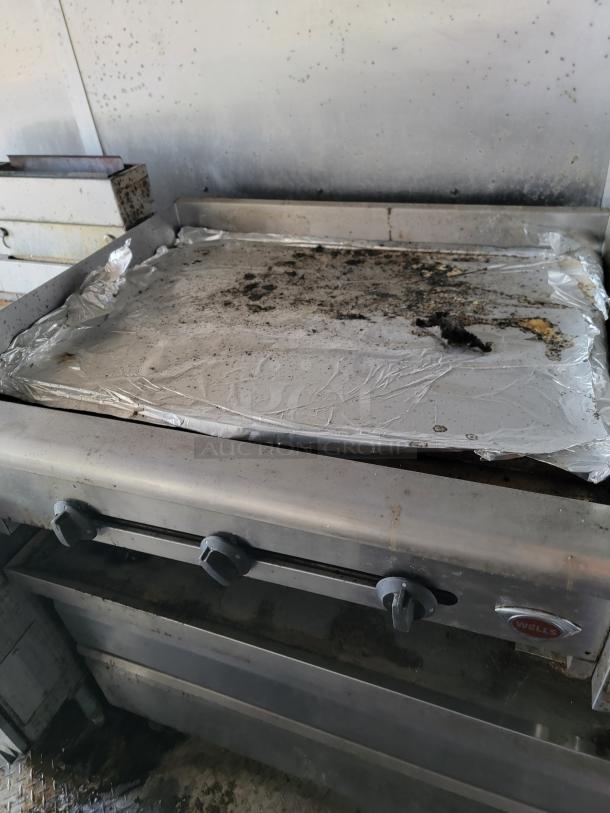 Commercial griddle by Wells, showing moderate wear, aluminum foil covered surface with residue, three control knobs visible.