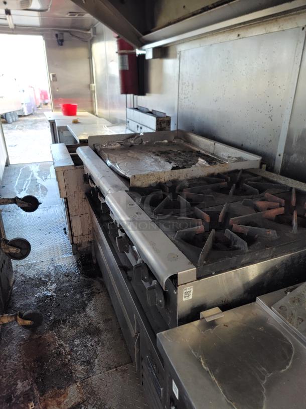Commercial kitchen stove with six burners and flat griddle, stainless steel, signs of use, located in a mobile food truck.