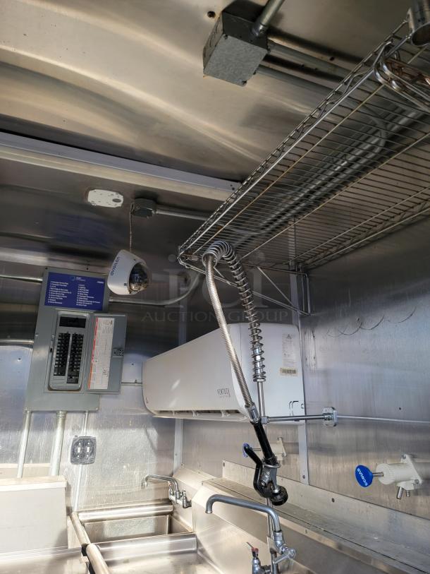 Commercial kitchen setup with stainless steel double sink, pre-rinse faucet, and overhead wire shelf. Includes Daikin air conditioning unit and circuit breaker panel, all in good condition.
