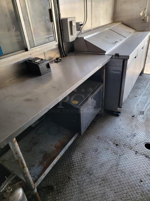 Stainless steel commercial kitchen prep area with refrigerator units, POS terminal, and storage shelf. Shows signs of rust.