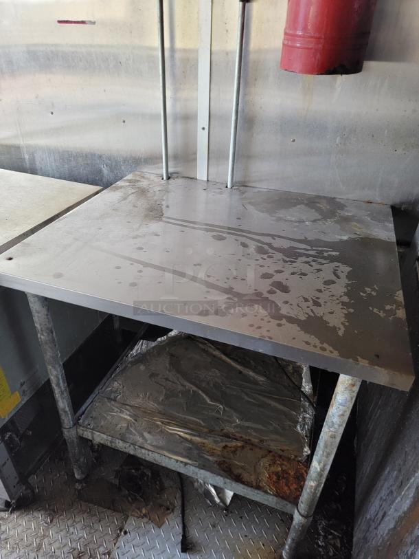 Stainless steel worktable with lower shelf, visible signs of wear and rust, sturdy metal legs, suitable for industrial use.