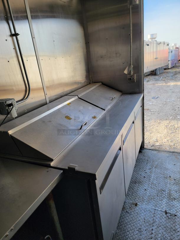 Commercial stainless steel kitchen counter with storage, showing moderate wear. Wire conduits and outlets visible on the wall.