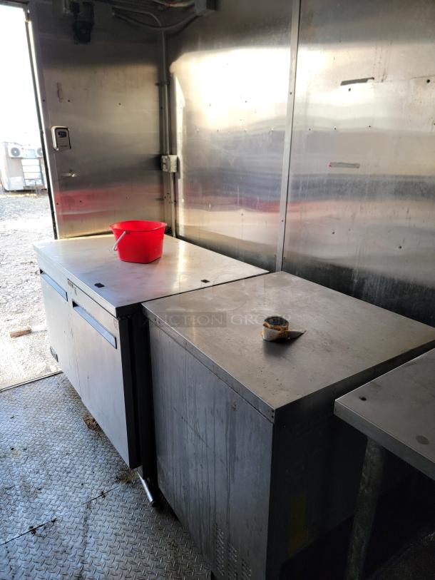Commercial stainless steel kitchen counters with storage space, featuring visible wear. Includes a red bucket and measuring tape on top.